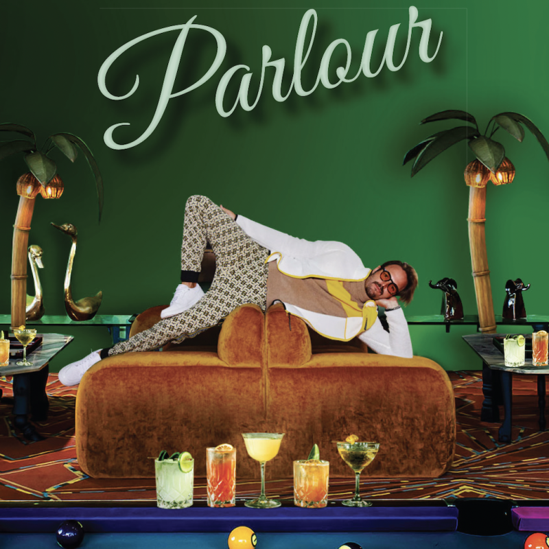 Man posing on couches behind a row of cocktails placed on a pool table.